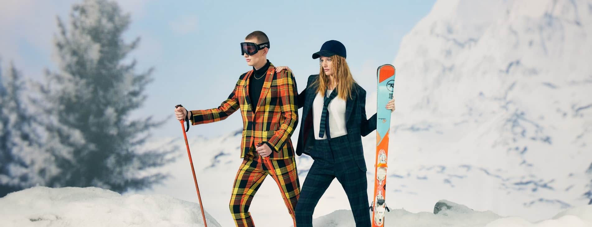 man in checked suit holds ski poles next to a women in a suit holding skis