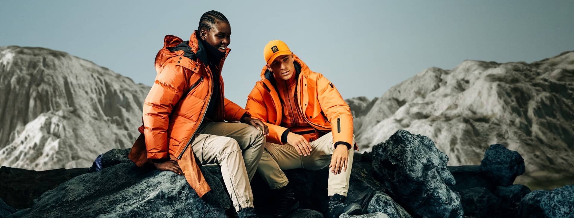 a woman and a man sat on a mountain range in orange Belstaff jackets